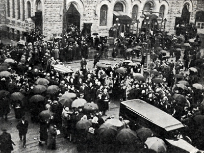 black and white image of a crowd of people, many holding large black umbrellas, in the street outside of a church, standing to the sides as a funeral procession moves through the square