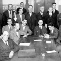 a large group of men and one woman sitting and standing around a long table, posing for a group photo